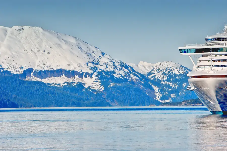 Alaska cruise ship bow navigating calm fjord waters with snow-capped forested mountains in the background.