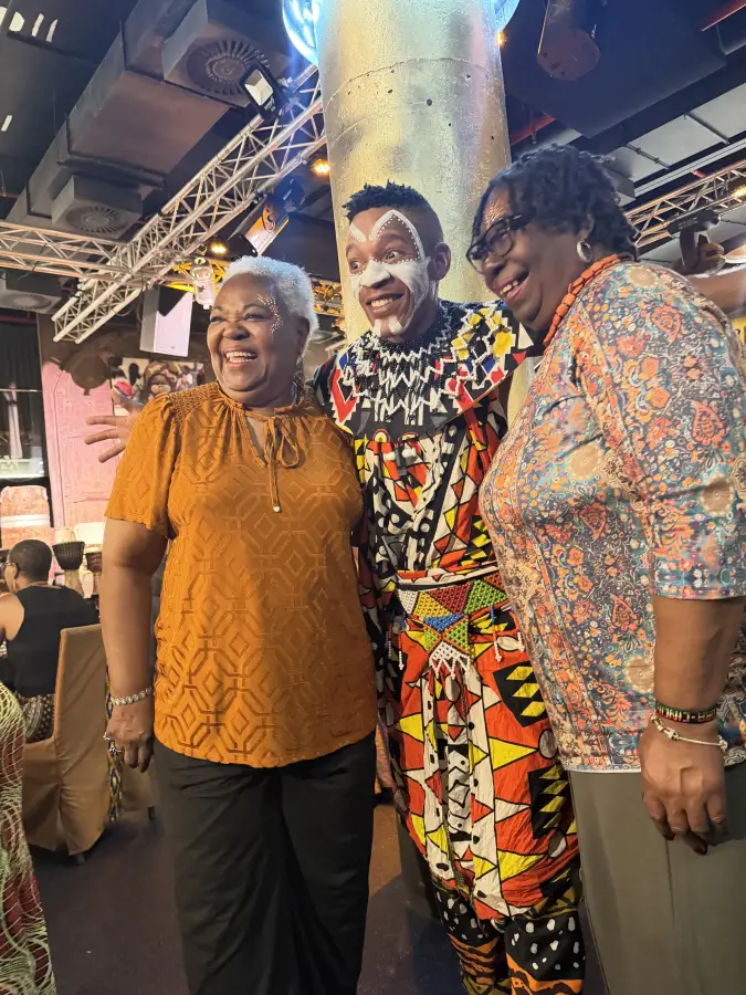 Two smiling women pose with a cultural performer in traditional face paint and beaded attire at the eat-mea-2026 event.