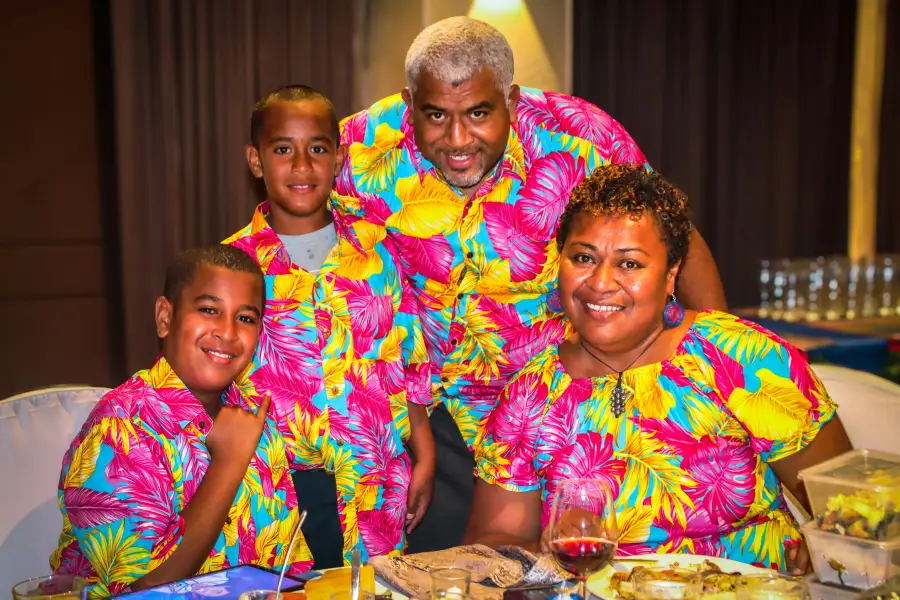 A smiling family of four wearing matching tropical print shirts enjoy a cruise dinner together at a festive table.