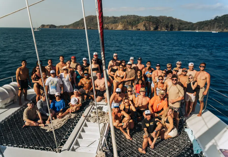 Large group of travelers posing together on a catamaran cruise deck with tropical island coastline in the background.