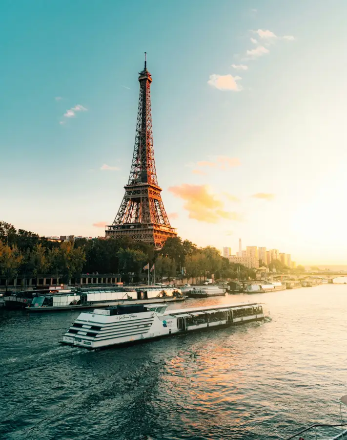 Paris Seine River cruise boat passing the Eiffel Tower at golden sunset; with city skyline in background.