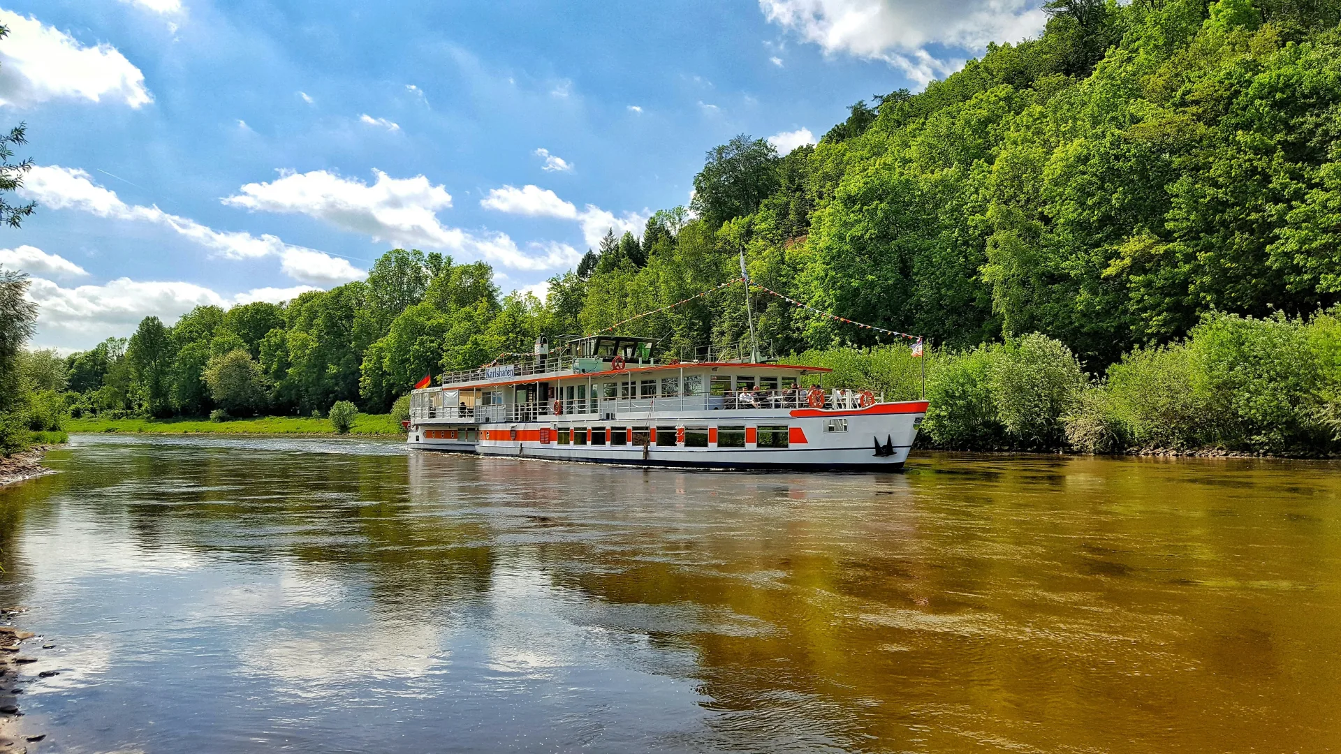 White and red river cruise vessel sailing along a calm tree-lined river under a partly cloudy summer sky.
