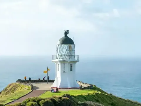 A bright white lighthouse stands on a lush green headland overlooking the vast blue ocean under a clear sky