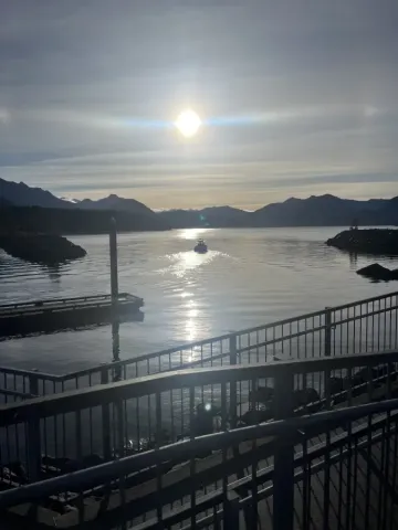A boat glides through calm Alaska Sitka harbor waters at sunset; with mountain silhouettes and a glowing sun reflected on