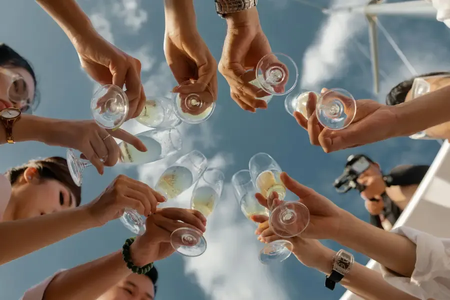 Group of guests raising champagne glasses in a circle toast on a cruise; photographed from below against a blue sky.