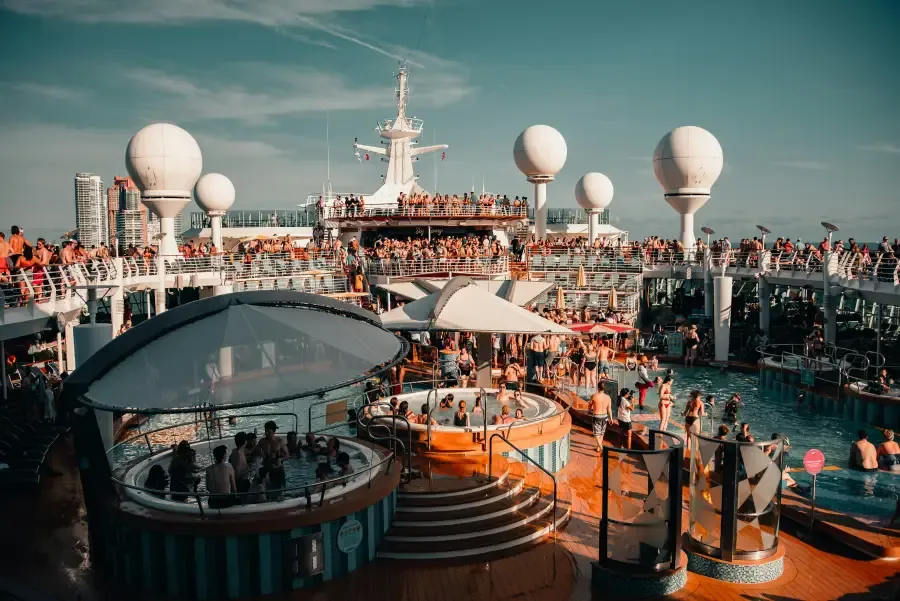 Crowded cruise ship pool deck with hot tubs; passengers; and white radar domes under a sunny sky near a city skyline.