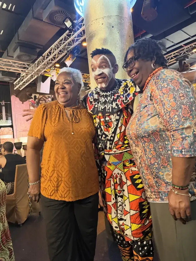 Two smiling women pose with a cultural performer in traditional face paint and beaded attire at the eat-mea-2026 event.