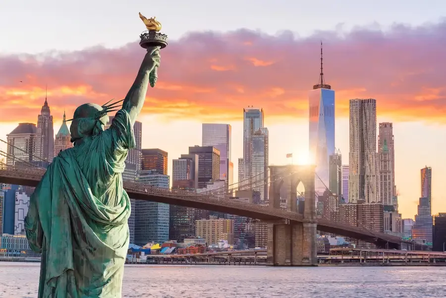 Statue of Liberty faces the New York New York skyline at sunset with Brooklyn Bridge and One World Trade Center behind