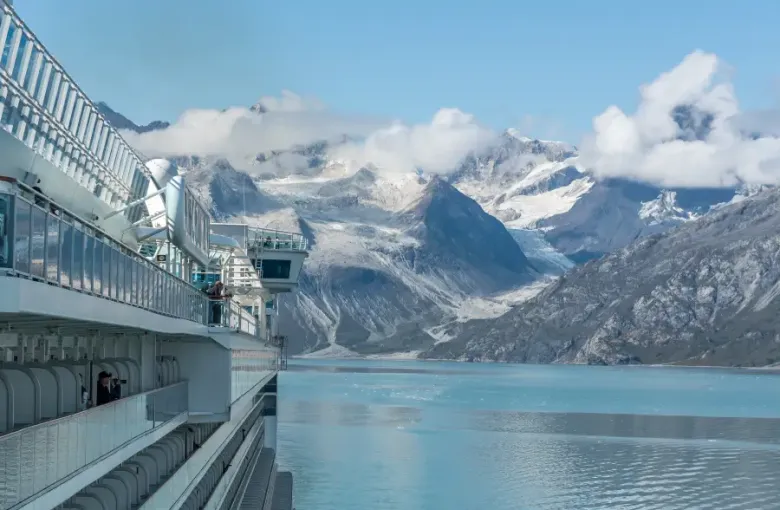Alaska cruise ship balconies overlooking glacial Glacier Bay waters with snow-capped mountains and flowing glaciers beyond.