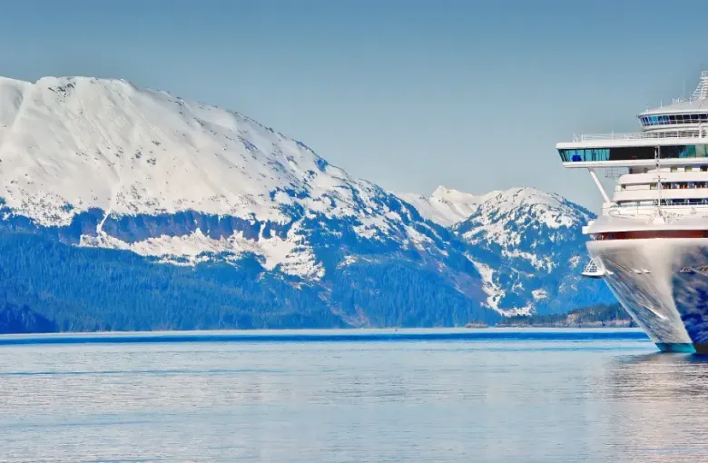 Alaska cruise ship bow navigating calm fjord waters with snow-capped forested mountains in the background.