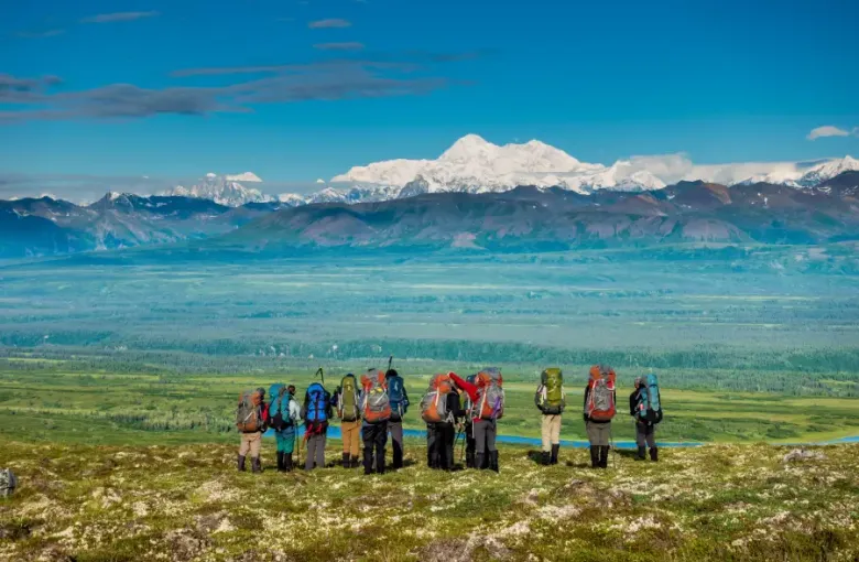 Group of backpackers standing on a tundra ridge overlooking a vast Alaska valley with Denali's snow-capped peak in the bac