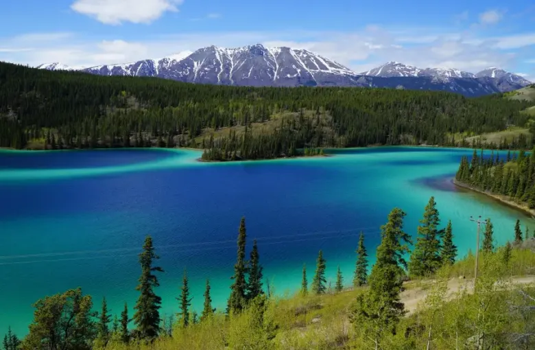 Vivid turquoise and deep blue glacial lake surrounded by boreal forest and snow-capped mountains in Alaska wilderness.