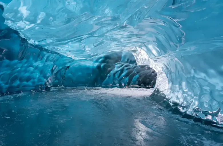 Glowing blue ice cave tunnel inside an Alaska glacier; with smooth frozen walls and icy floor leading to a dark passage.