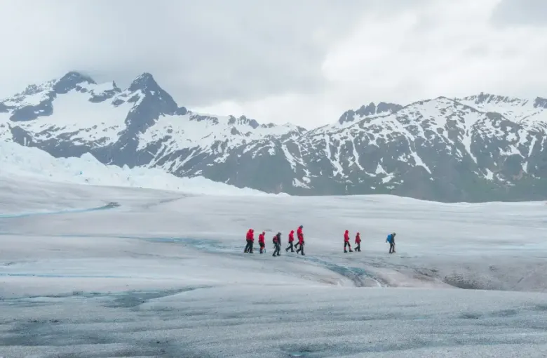 Group of hikers in red jackets trekking across an Alaska glacier with snow-capped mountains in the background.