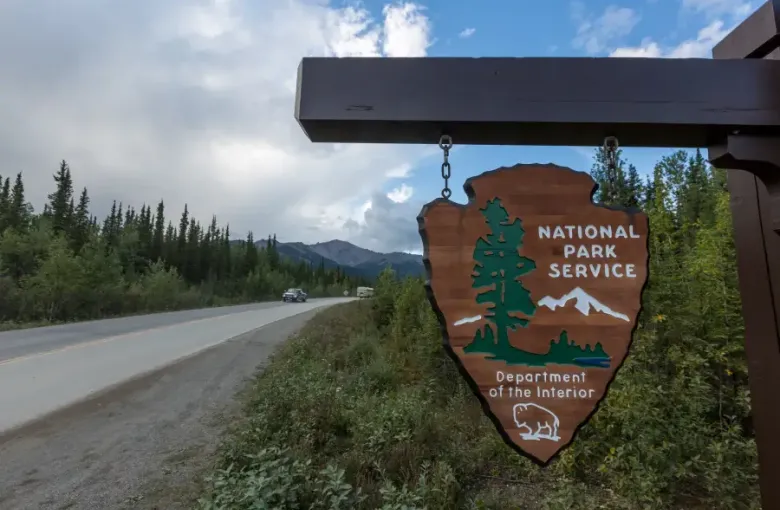 National Park Service arrowhead sign hanging roadside in Alaska; with forested highway and mountain range in background.