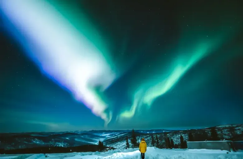 Traveler in yellow coat gazing at vivid green and white aurora borealis over snow-covered Alaska mountains at night.