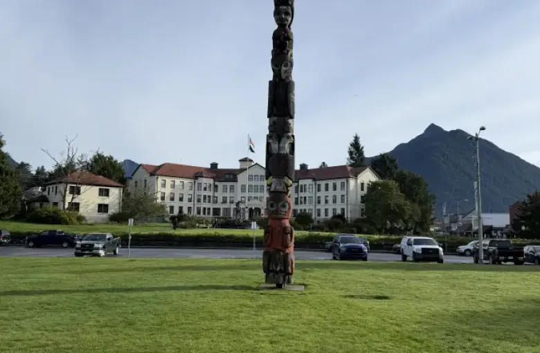 Tall carved totem pole standing on a green lawn in alaska-sitka; with a historic building and mountain in the background.