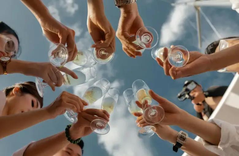 Group of guests raising champagne glasses in a circle toast on a cruise; photographed from below against a blue sky.