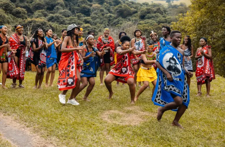 Group of young adults in vibrant traditional African attire performing a cultural dance outdoors on green grass; cruise-la