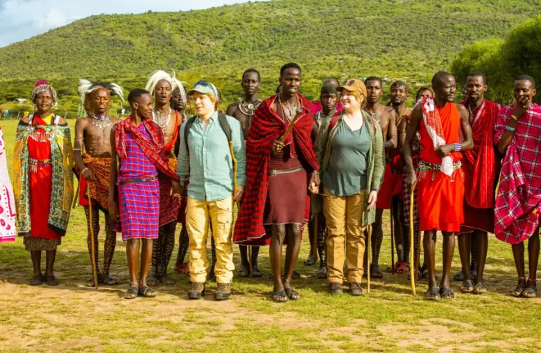 Two Western tourists stand among traditionally dressed Maasai community members during a cruise-land excursion in Kenya's