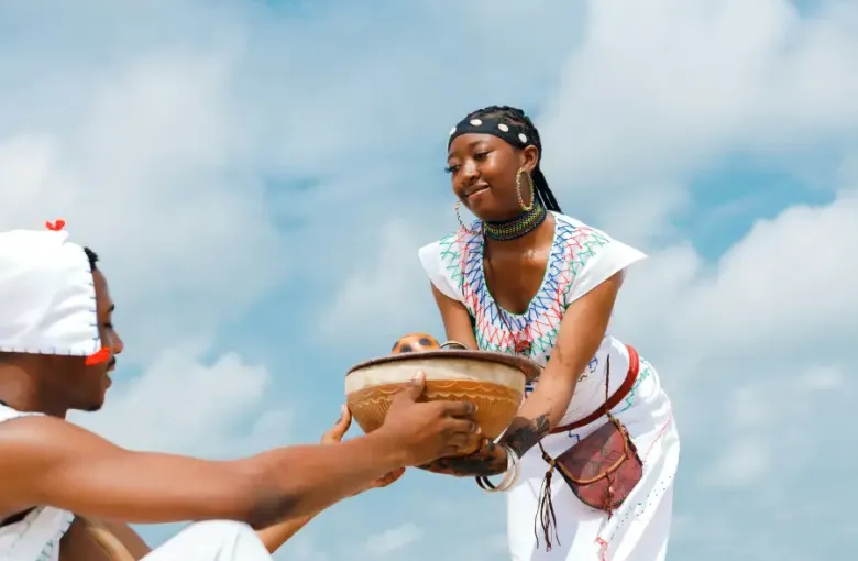Woman in traditional African attire with beaded jewelry offers a carved wooden bowl during a cruise-land cultural ceremony.