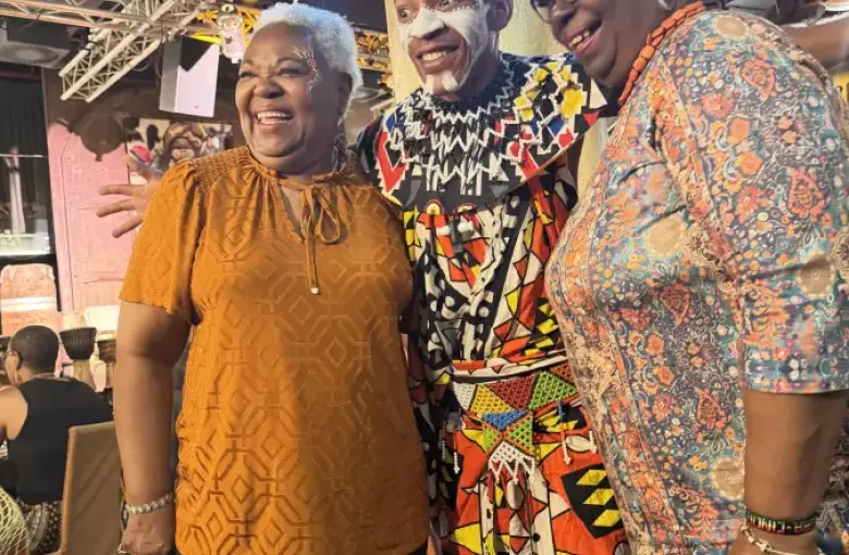 Two smiling women pose with a cultural performer in traditional face paint and beaded attire at the eat-mea-2026 event.
