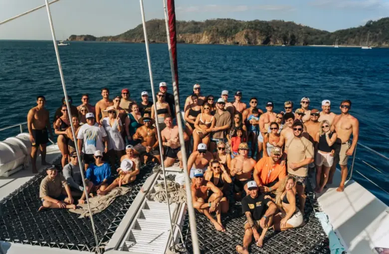 Large group of travelers posing together on a catamaran cruise deck with tropical island coastline in the background.