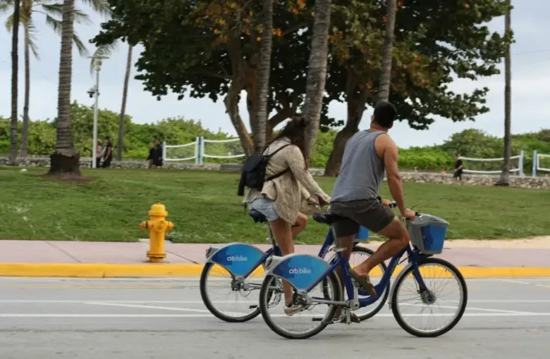 Couple riding Citi Bike rental bicycles along a palm-lined street in Miami; Florida on a cloudy day.