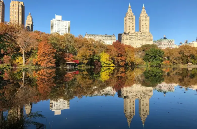 Central Park lake reflecting autumn foliage and Upper West Side skyscrapers in New York New York under a clear blue sky