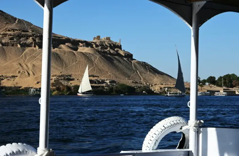 River cruise view of feluccas sailing the Nile near Aswan's ancient hillside tombs under clear blue sky.