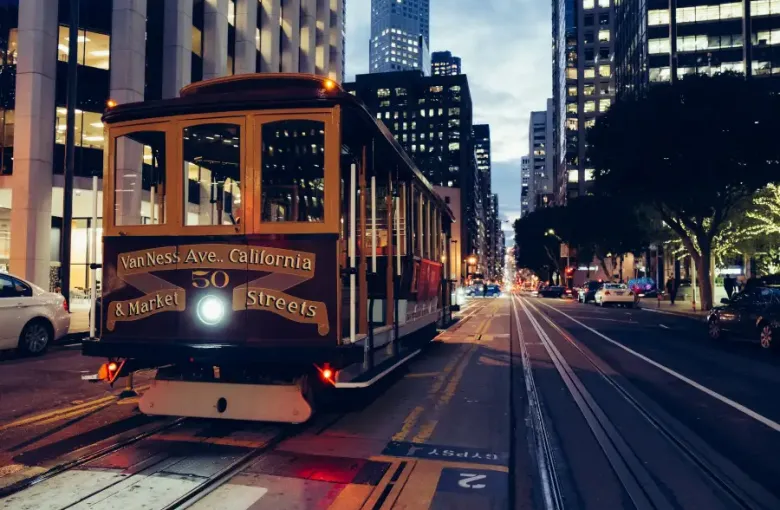 San Francisco CA cable car number 50 on California Street at dusk; with downtown skyscrapers and city lights in background.