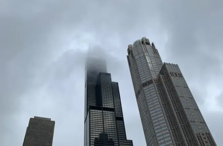 Willis Tower piercing low clouds on a stormy day in Chicago; viewed from street level alongside neighboring skyscrapers.
