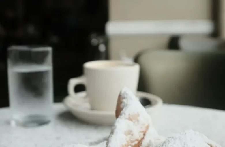 A plate of warm New Orleans beignets covered in powdered sugar with a cup of cafe au lait