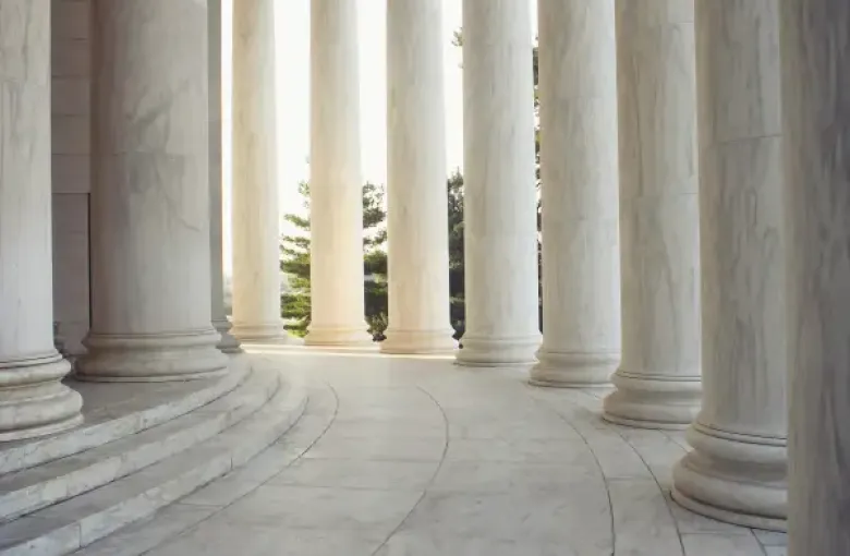 Curved marble colonnade of the Jefferson Memorial in Washington; DC; with Ionic columns framing sunlit trees beyond.