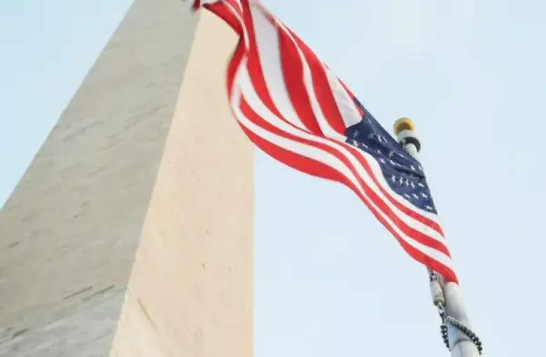 American flag waving beside the Washington DC Monument obelisk viewed from below against a clear blue sky.