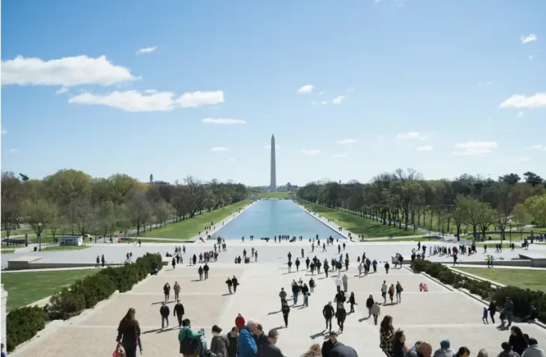 Tourists walk along the National Mall in Washington DC with the Reflecting Pool and Washington Monument in the background.